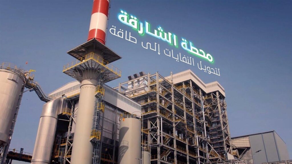 The Sharjah Waste-to-Energy Plant, a sleek, modern facility symbolizing the UAE’s commitment to sustainability. The plant’s clean architectural lines and tall emission stack stand against a bright blue sky, representing innovation and environmental progress. In the foreground, solar panels and landscaped greenery highlight the integration of technology and nature, a visual statement of the UAE’s journey toward a zero-waste future.